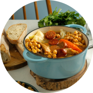 Refeição caseira de grão de bico, legumes e chouriço, servida numa panela azul, juntamente com fatias de pão numa mesa de madeira